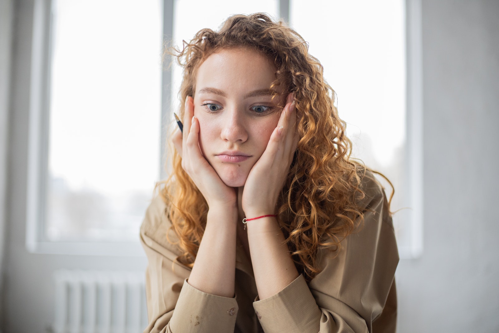 Pensive female student with hands on cheeks looking down while sitting in light room on blurred background while deciding task in frustration