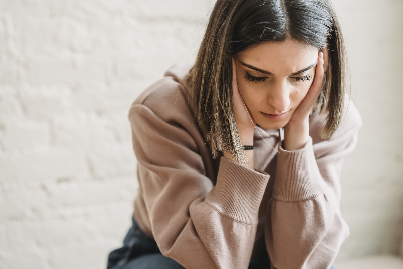 Crop unhappy female in casual wear touching cheeks and looking down while sitting near white wall in light room at home