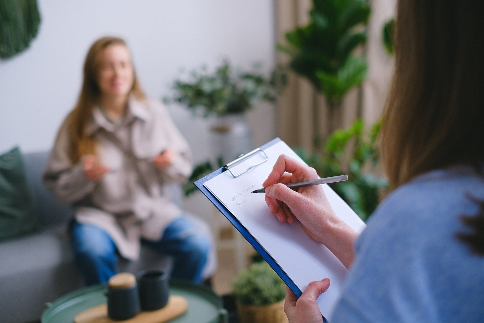 Unrecognizable professional female psychologist writing on clipboard while sitting against client on blurred background during psychotherapy session in light office