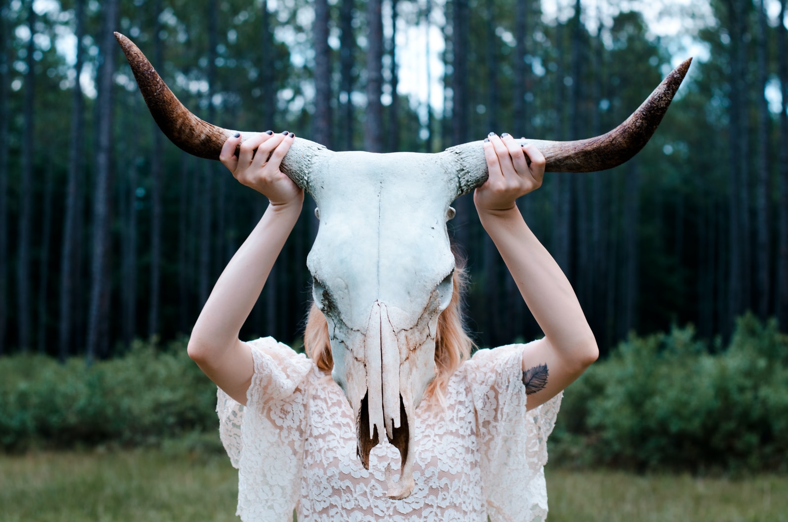 woman holding white and black animal skull wearing white lace floral sleeveless dress near green leaf trees