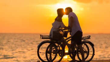 Photography of Man Wearing White T-shirt Kissing a Woman While Holding Bicycle on River Dock during Sunset