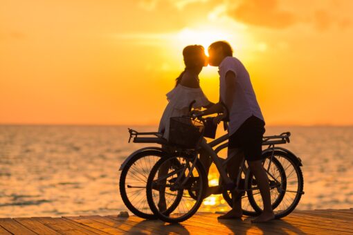 Photography of Man Wearing White T-shirt Kissing a Woman While Holding Bicycle on River Dock during Sunset