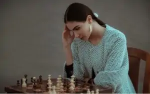 Concentrated ethnic female sitting on chair and touching head while playing chess against blurred gray background
