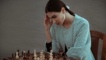Concentrated ethnic female sitting on chair and touching head while playing chess against blurred gray background