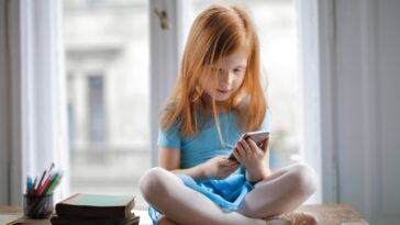 Red haired charming schoolgirl in blue dress browsing smartphone while sitting on rustic wooden table with legs crossed beside books against big window at home