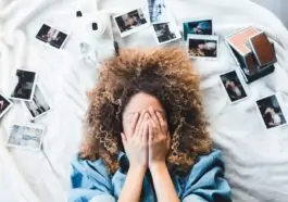 woman lying on bed covering her face surrounded by photos and white camera