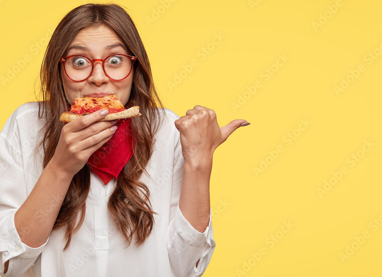 Lunch time concept. Attractive cheerful woman eats delicious Italian pizza, surprised with wonderful taste, indicates with thumb aside, shows where pizzeria situated, suggests to visit and have snack