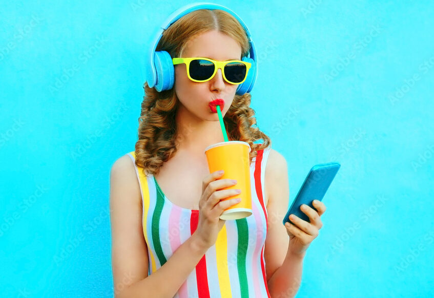 Portrait cool girl drinking fruit juice holding phone listening to music in wireless headphones on colorful blue background